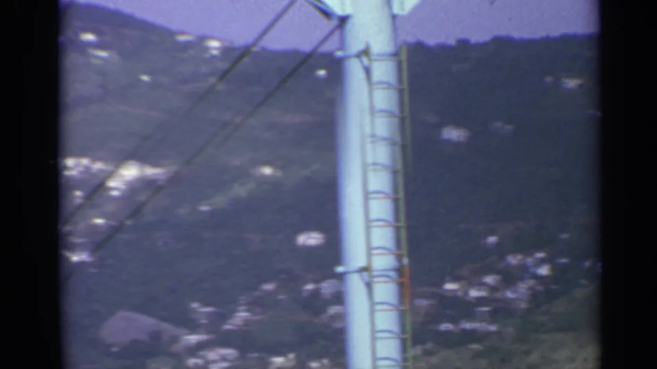 A pale utility pole with a ladder dominates the foreground, with a wooded US Virgin Islands hillside and small buildings behind it.