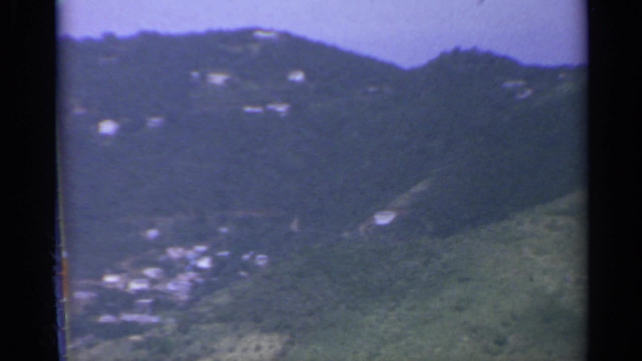 Green hillside with scattered pale buildings seen from the air, bordered by dark film edges.