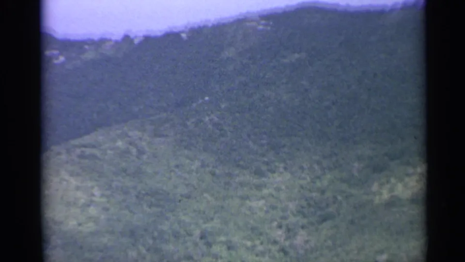 Aerial 8mm view of a tropical hillside, with dense vegetation and a small distant building cluster on the slope.