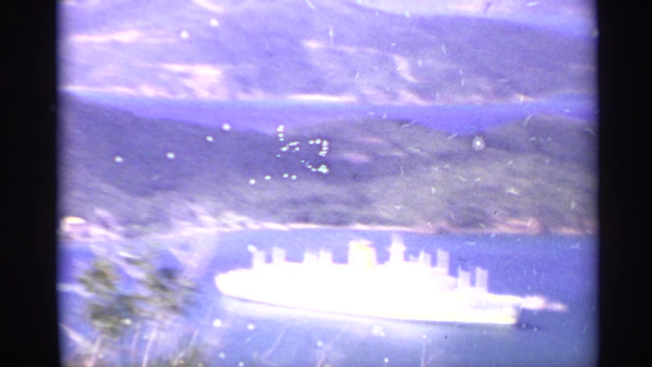 Large white passenger ship on blue water below mountain scenery.