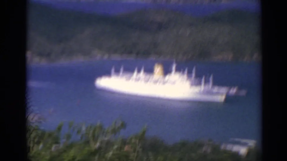 1967 US Virgin Islands frame with the ship softened by camera movement, framed by foliage and mountain shoreline.