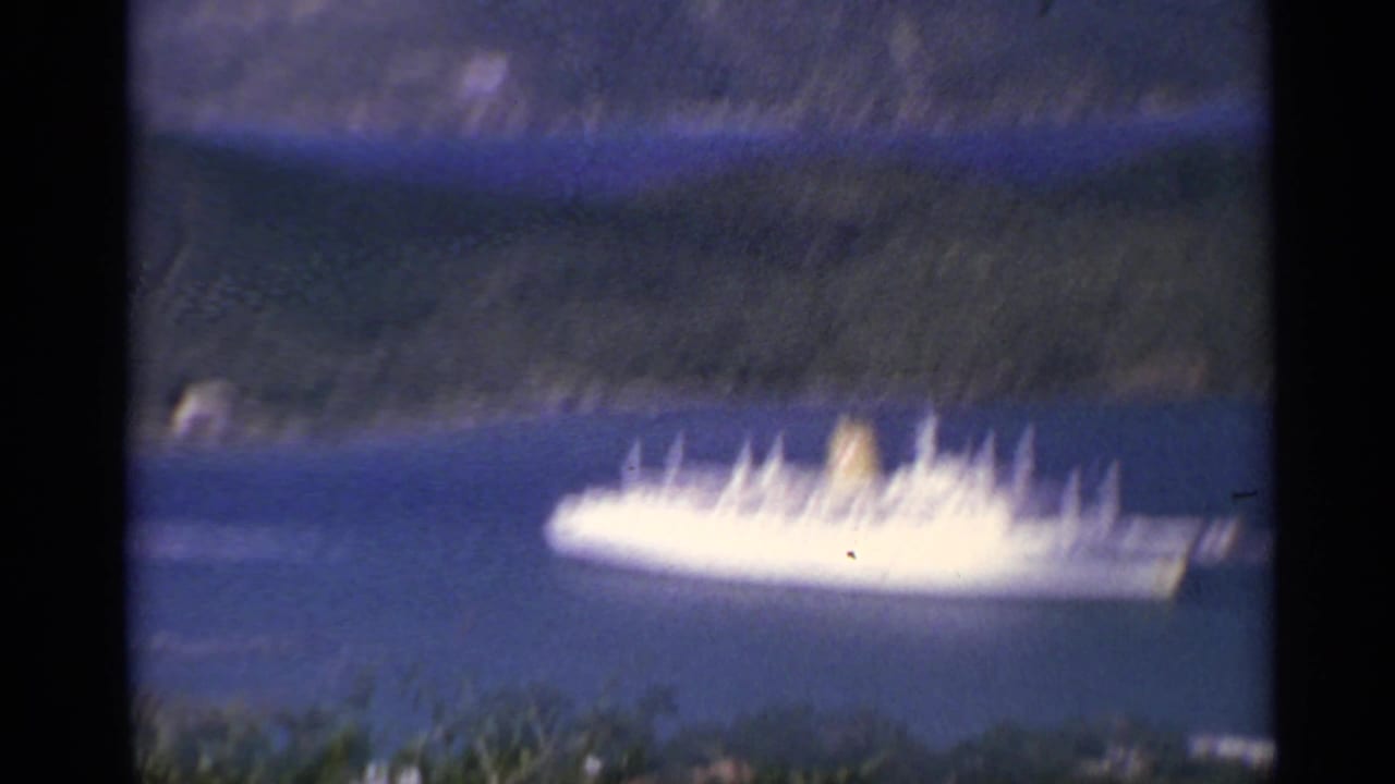 1967 8mm travel frame showing a white cruise ship on deep blue water with green mountain slopes behind it.