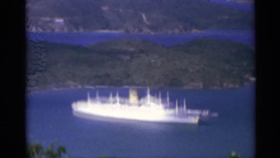 White passenger ship seen from above in a blue bay, with wooded island hills and shoreline in the background.