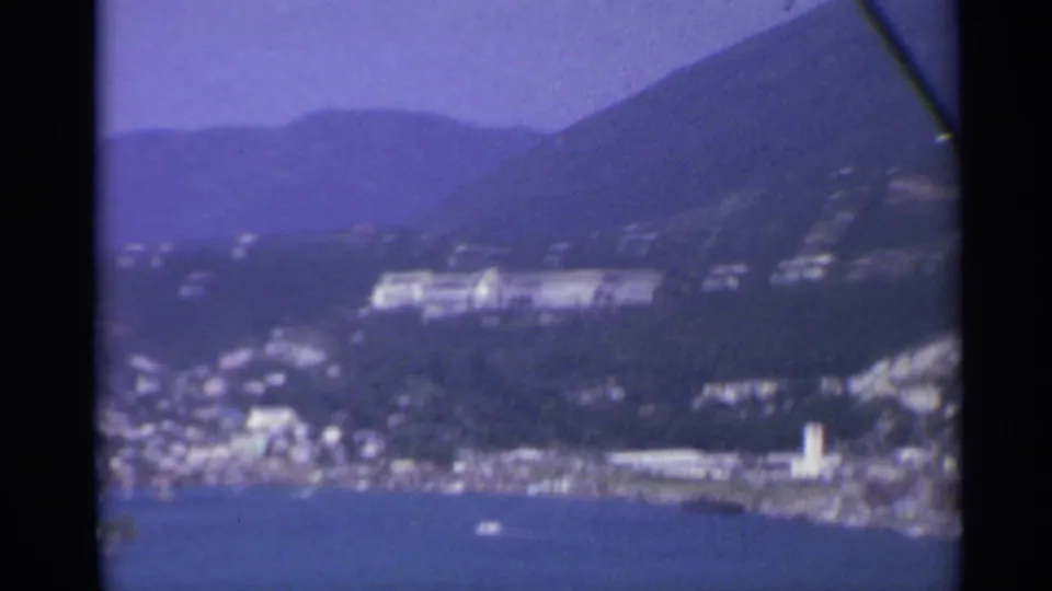 Soft 8mm frame of a harbor shoreline with white buildings, blue water, and steep mountain slopes beyond.