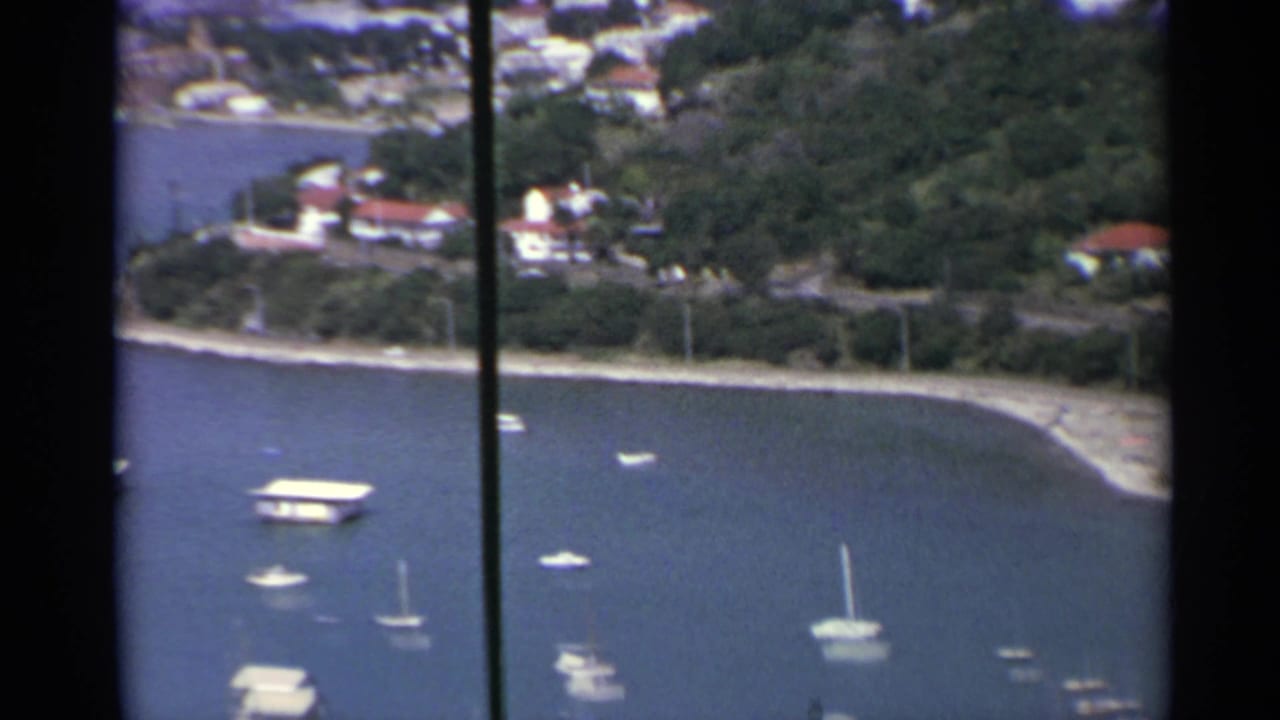 Elevated harbor view with blue water, anchored boats, shoreline, and hillside buildings.