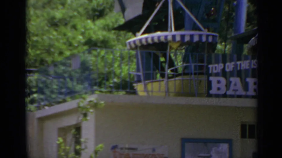 1967 visitor-shot frame showing greenery, a balcony structure, and a partial readable Top of the Island Bar sign.