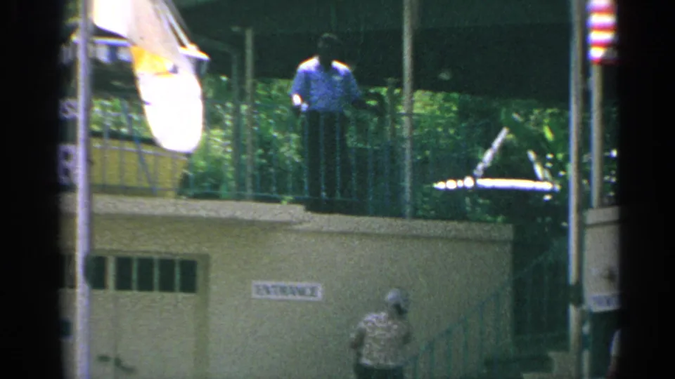 1967 US Virgin Islands home-movie frame showing a man on the balcony, flags in the foreground, stairs, and a person below near the facade.