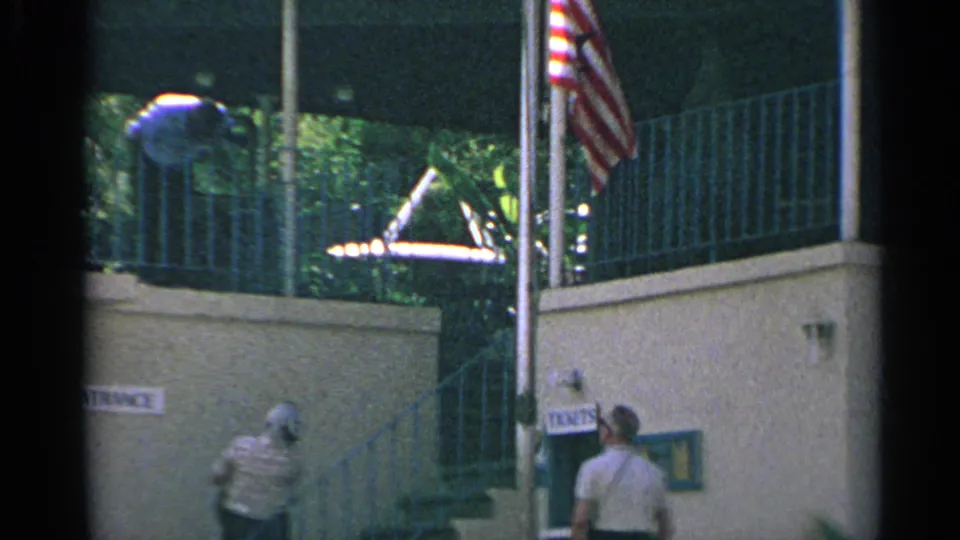 1967 US Virgin Islands frame with a balcony, American flag, exterior stairs, one person leaning above, and another person walking below.