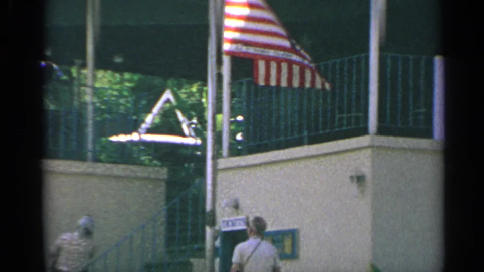 1967 8mm US Virgin Islands travel frame showing an American flag, tramway station facade, ticket booth, and visitors below.