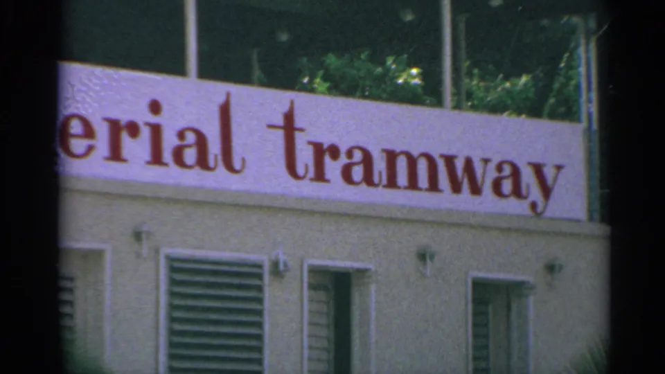 1967 8mm US Virgin Islands frame with the tramway sign continuing across the station facade and greenery above.