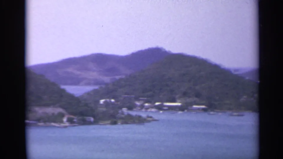 1967 8mm island panorama with green hills, blue water, and distant shoreline structures.