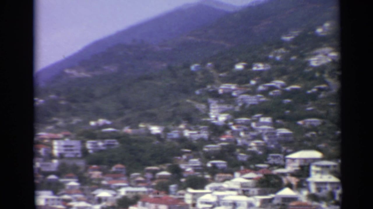 1967 US Virgin Islands home-movie view of houses clustered on a green hillside beneath hazy mountain ridges.