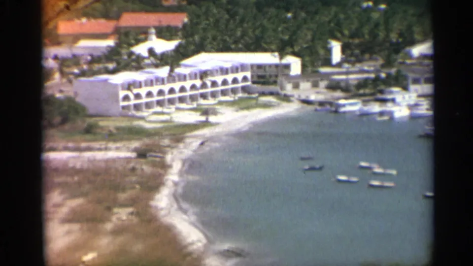 1967 US Virgin Islands harbor view with a curved beach, white arched waterfront building, and small boats on calm water.