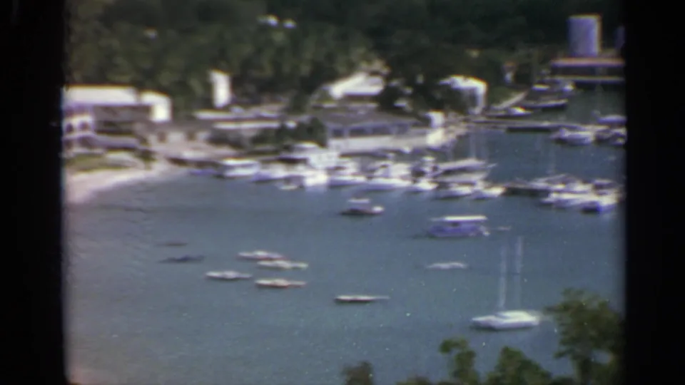 1967 US Virgin Islands harbor with anchored boats, shoreline structures, green hillside, and a distant water tank.
