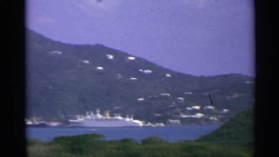 1967 US Virgin Islands harbor scene with a distant cruise ship, hillside homes, and foreground vegetation.