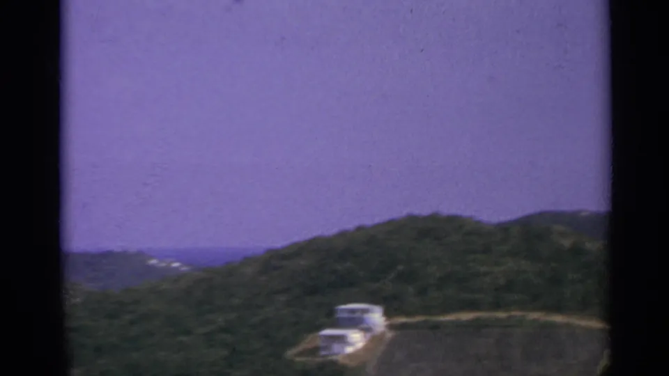 Elevated US Virgin Islands view with a hilltop road, white buildings, green slopes, and distant coastal water in soft 8mm color.