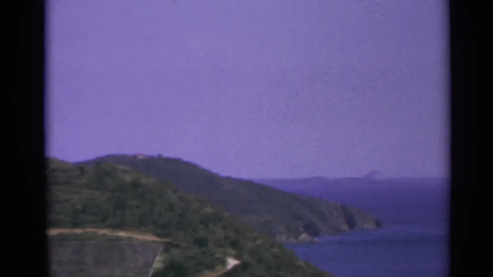 1967 US Virgin Islands home-movie frame of a green coastal peninsula, curving shoreline, blue water, and purple-tinted sky.