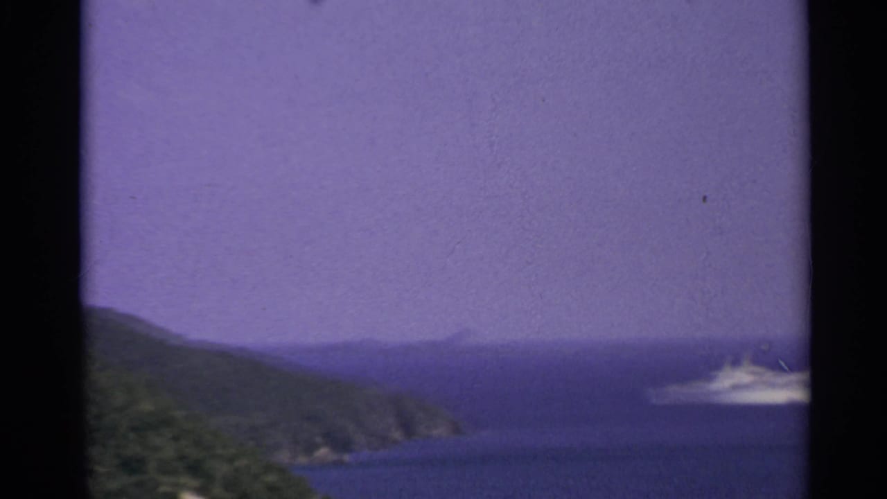 White ship on blue water near green coastal hills and a hazy sky.