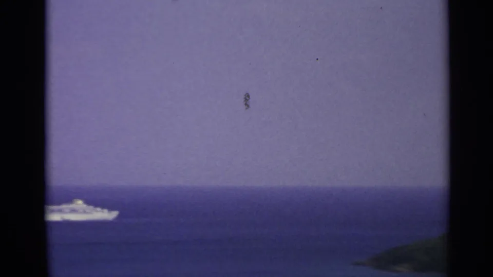 A white cruise ship slips partly out of frame in a 1967 8mm coastal view of blue sea and green land.