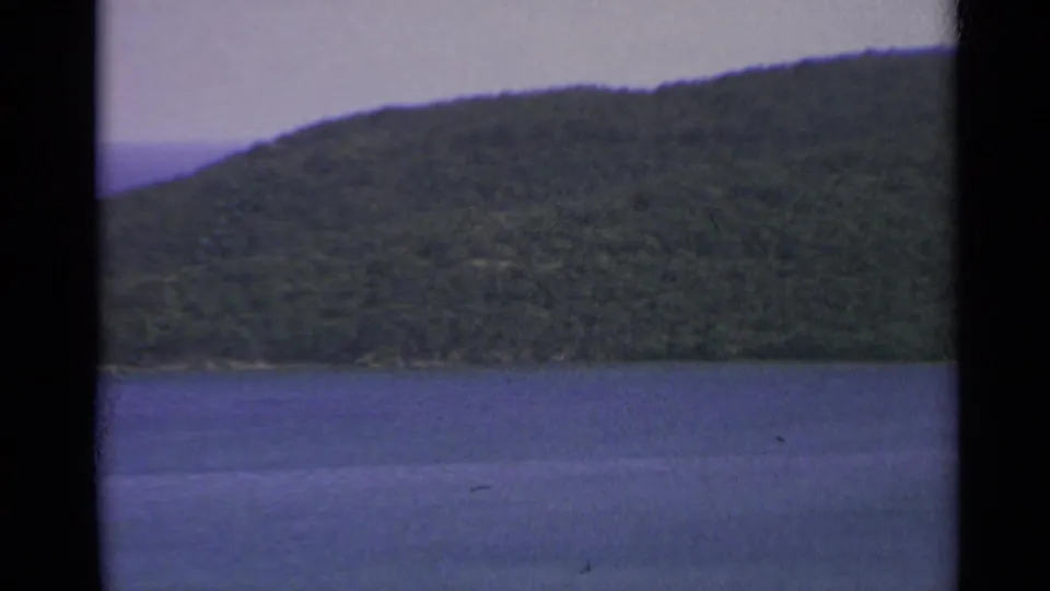 1967 US Virgin Islands frame showing blue coastal water below a forested hillside and distant shoreline.