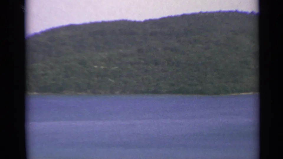 1967 US Virgin Islands 8mm view across blue water toward a steep green hillside and narrow shoreline.