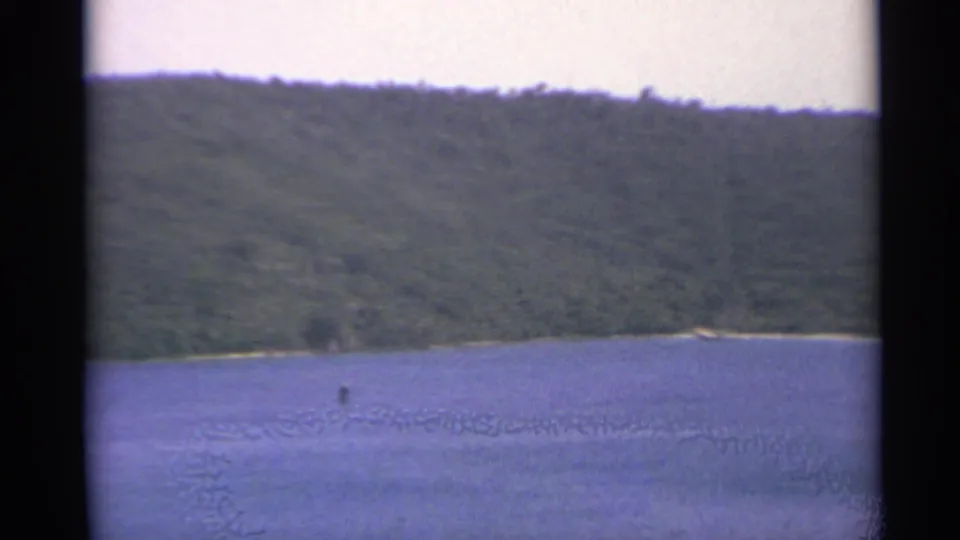 1967 US Virgin Islands 8mm frame showing water, a forested hillside, pale sand, and a distant small boat or person.