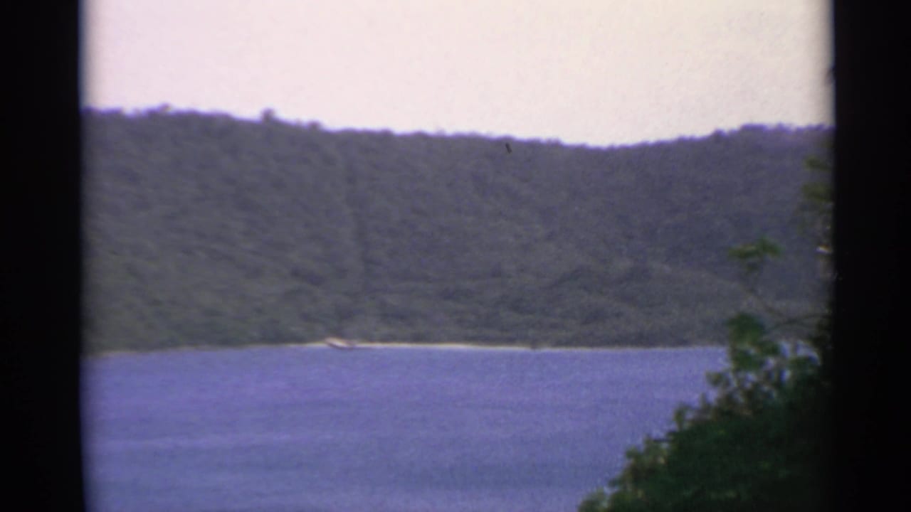 Blue water below forested hills with pale sky and dark film borders.