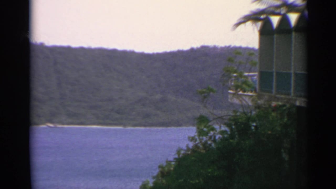 1967 US Virgin Islands landscape with blue water, green hills, palm foliage, and a partial arched building at right.
