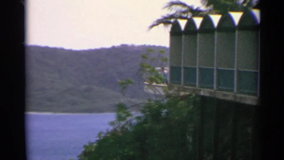 1967 US Virgin Islands 8mm frame showing an arched green-and-white building, foliage, blue water, and wooded hills.