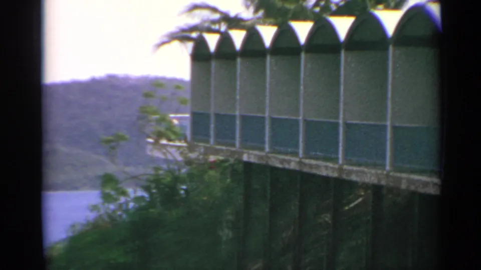 1967 US Virgin Islands frame with a green-and-white arched building, palm fronds, dense greenery, water, and distant hills.