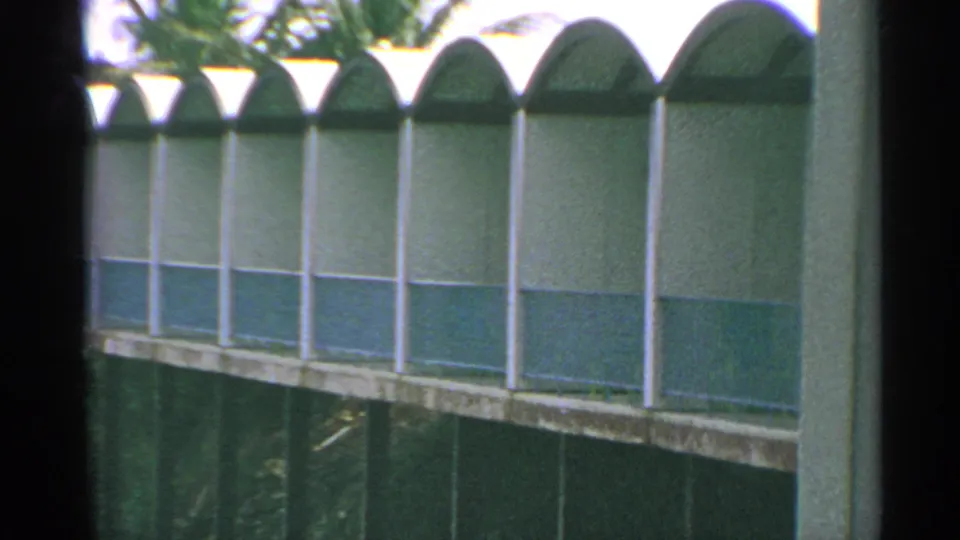 The camera frames the arched coastal building more centrally, showing repeated window bays and tropical foliage behind it.
