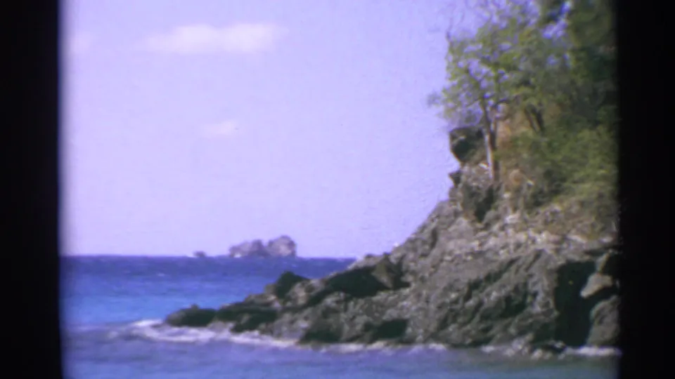 1967 US Virgin Islands coastal view showing a rocky headland, blue ocean, pale sky, and small offshore rock formation.