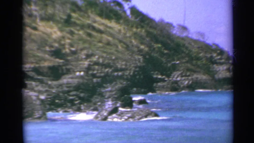 1967 US Virgin Islands 8mm frame with calm turquoise water, shoreline rocks, surf patches, and a steep green coastal hillside.