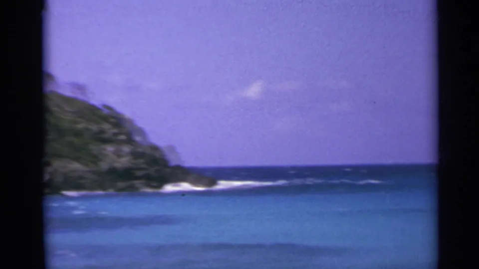 1967 US Virgin Islands 8mm view of open turquoise water, a rocky green headland, surf at the base, and hazy bright sky.