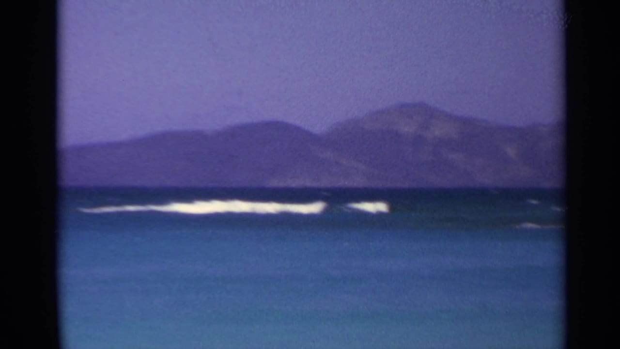 Turquoise bay with white surf, distant hills, and dark film-frame borders.