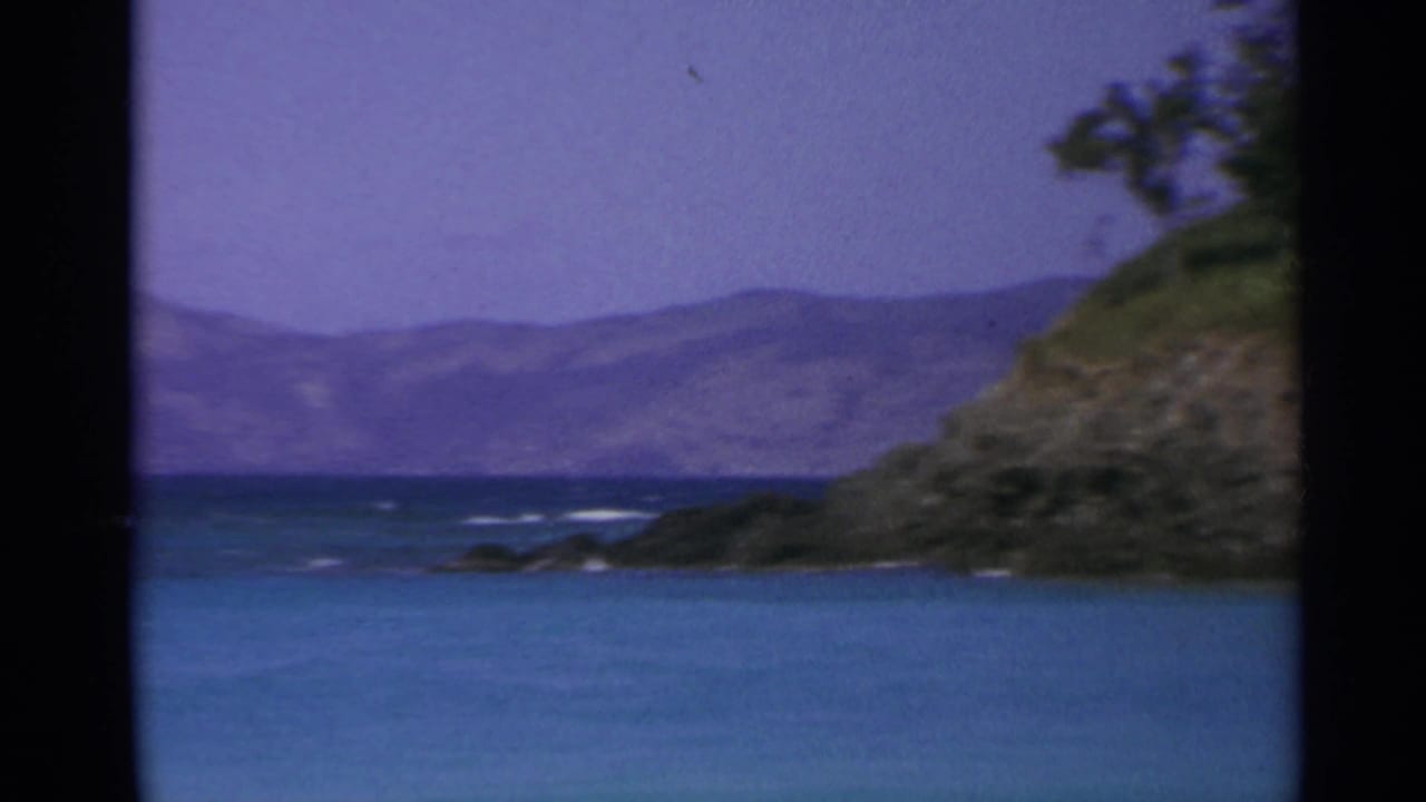 Rocky green shoreline beside calm blue water with distant hills beyond.