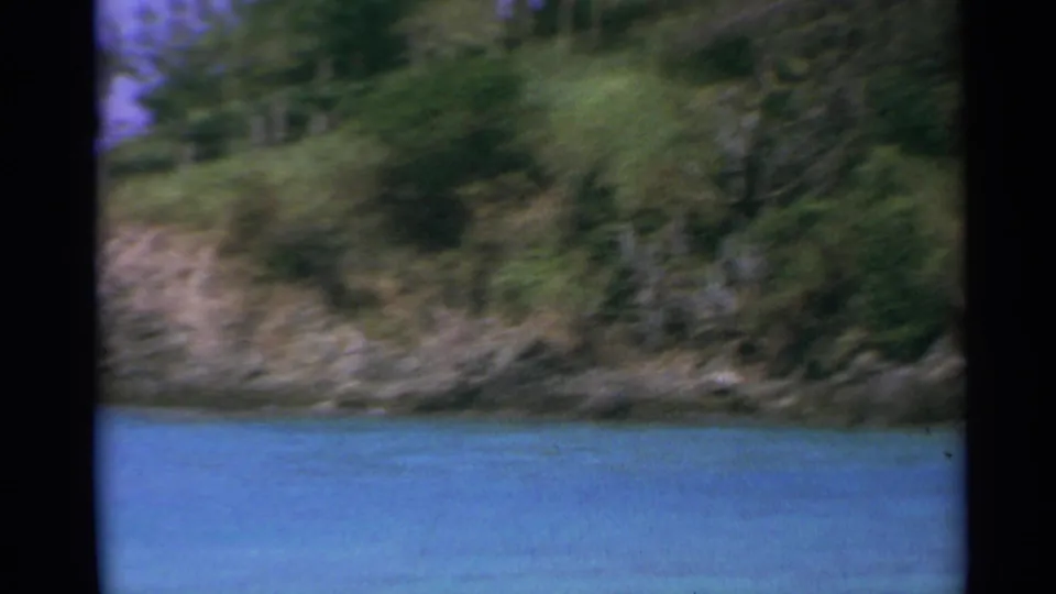 Handheld 1967 US Virgin Islands home-movie frame showing a green, rocky shoreline above clear blue water.
