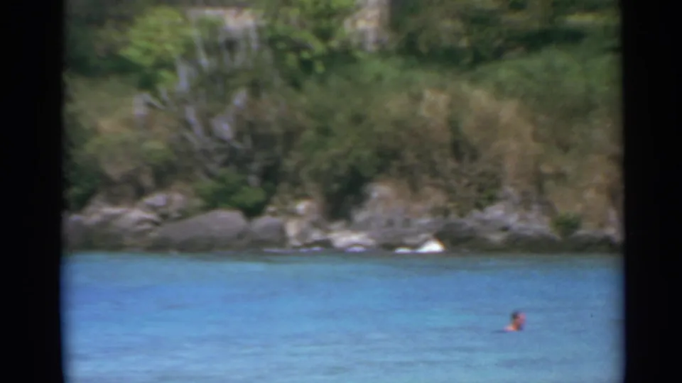 1967 US Virgin Islands home-movie frame with a distant swimmer in blue water below a rocky green shore.