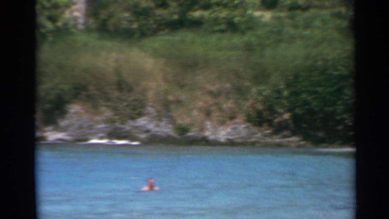 1967 US Virgin Islands home-movie frame showing a swimmer in turquoise water below a green, rocky shoreline.