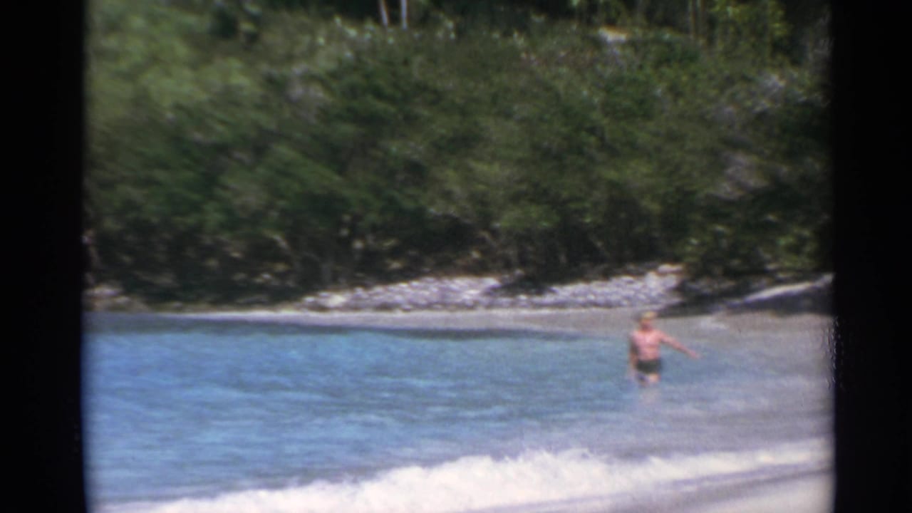 Adult man wading near the surf with vegetation and rocks behind the beach.