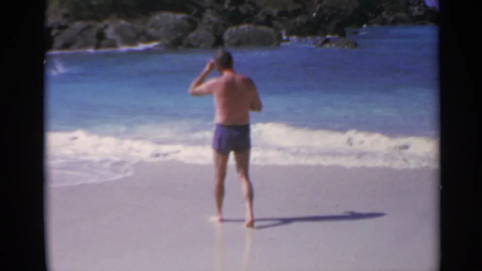 1967 US Virgin Islands home-movie frame showing a shirtless adult man on wet sand facing the ocean.