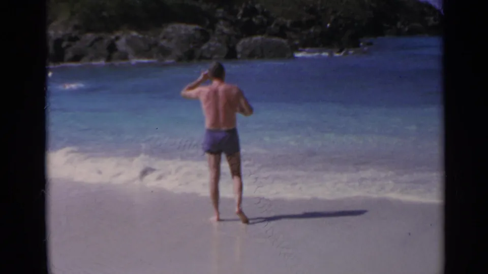 1967 US Virgin Islands beach frame with an adult man walking at the shoreline beside clear turquoise water.