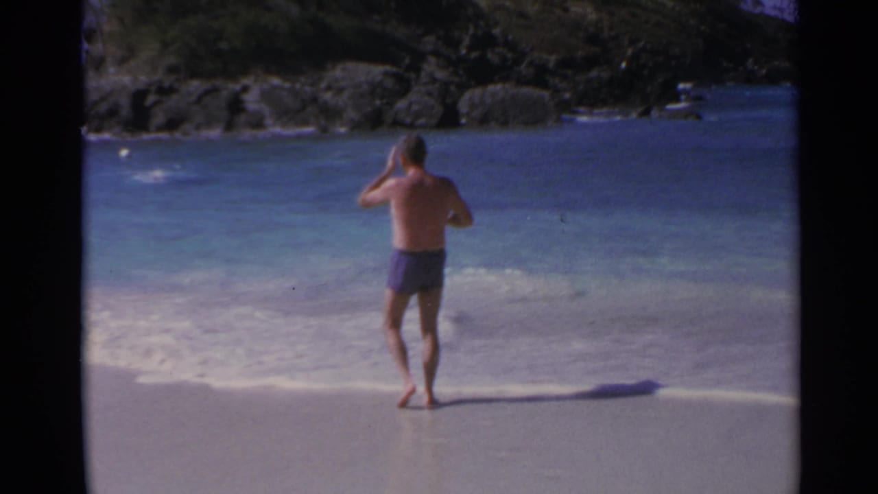1967 8mm home-movie frame of a man in purple swim trunks standing barefoot at a US Virgin Islands shoreline with rocks beyond.