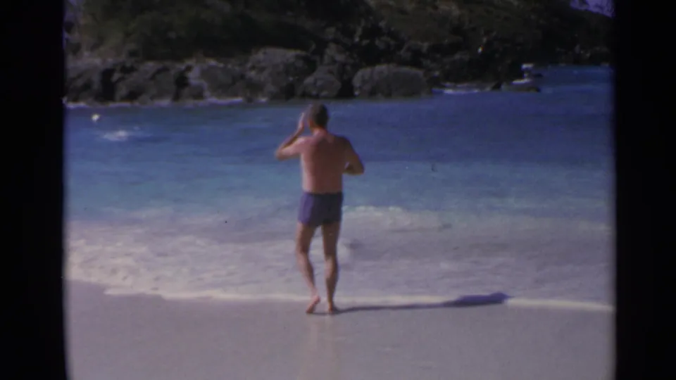 1967 8mm home-movie frame of a man in purple swim trunks standing barefoot at a US Virgin Islands shoreline with rocks beyond.