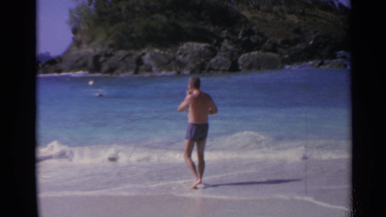 Man in swim trunks stands near a small breaking wave with rocky shoreline in the background.