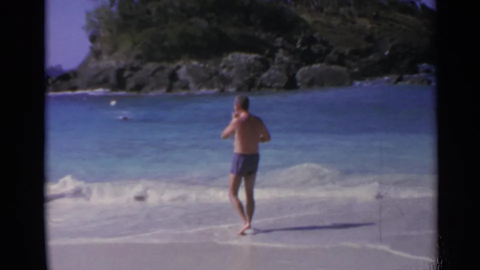 Man in purple trunks faces the turquoise water on a US Virgin Islands beach in a 1967 8mm vacation shot.