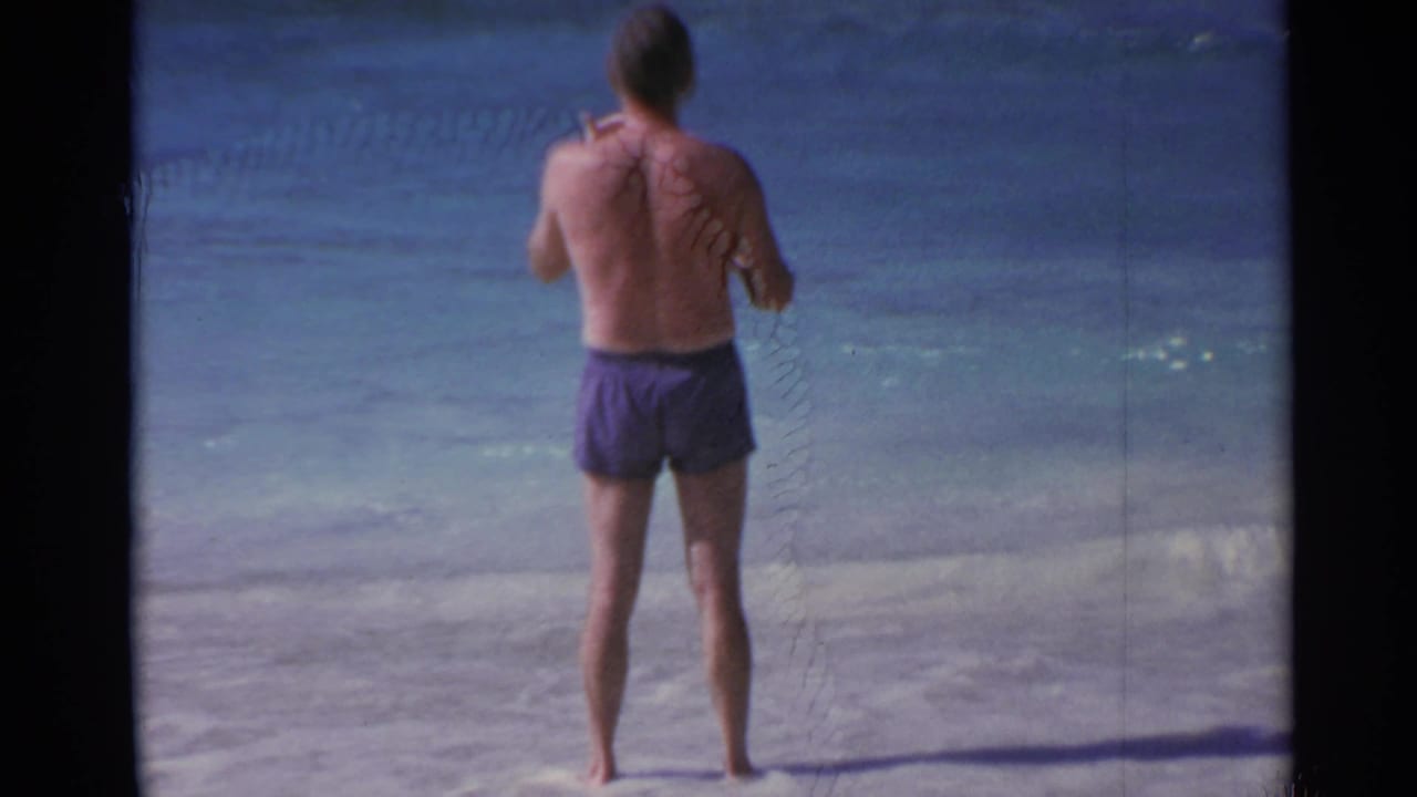 Barefoot man in purple swim trunks stands on a sandy beach facing the ocean.