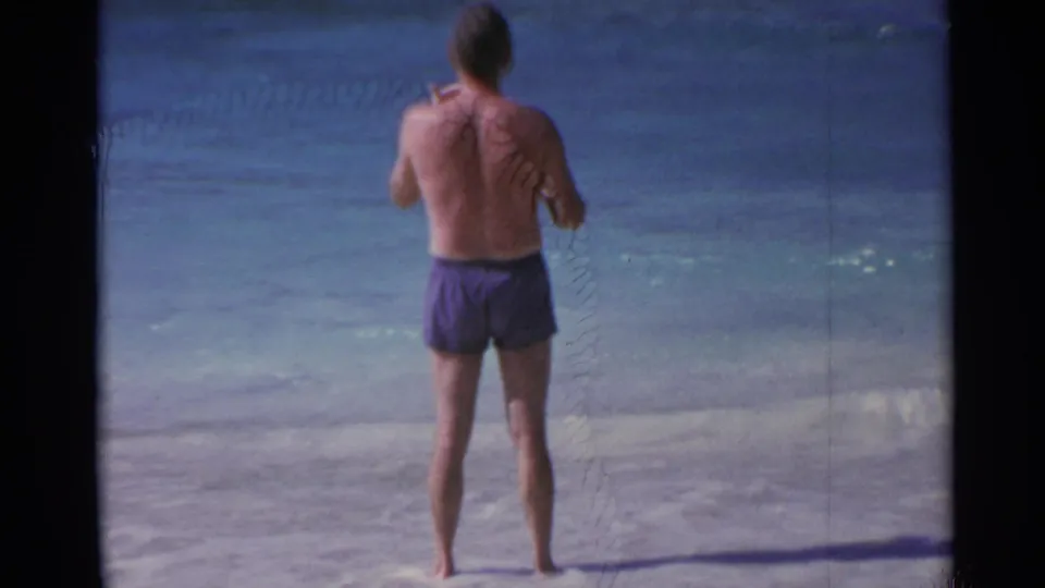 In 1967 U.S. Virgin Islands home-movie footage, a barefoot man in purple swim trunks looks toward blue ocean water from a sandy beach.