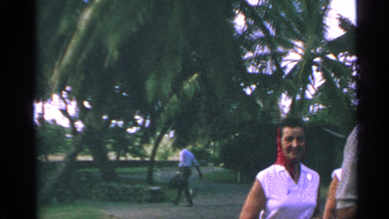 1967 US Virgin Islands home-movie frame with a woman in a white sleeveless top and red scarf walking past palm trees and a paved resort drive.