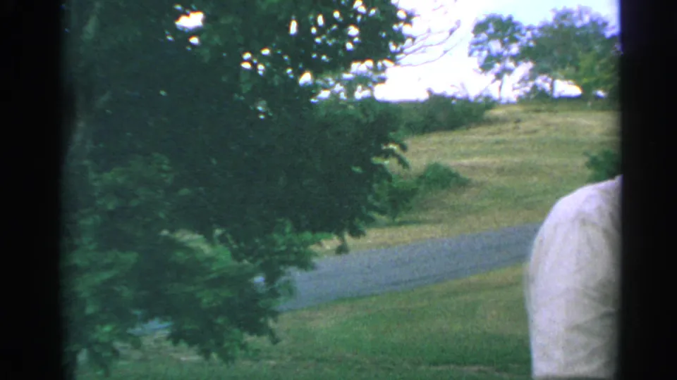 1967 US Virgin Islands home-movie frame with a partial shoulder or arm at right, foreground foliage, grass, and a curving path.
